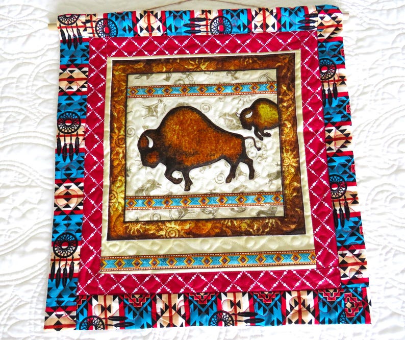 May include: A Native American-themed wall hanging featuring a central image of a brown bison. The artwork is framed with a red border and accented with turquoise and white geometric patterns. The piece is mounted on a wooden dowel.