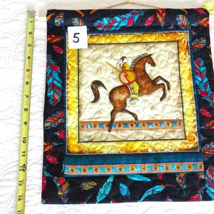 May include: Quilted wall hanging featuring a Native American on horseback. The central image is framed with a yellow border and a dark blue and turquoise border. The outer border is black with colorful feather designs. The wall hanging measures approximately 14 inches in length.