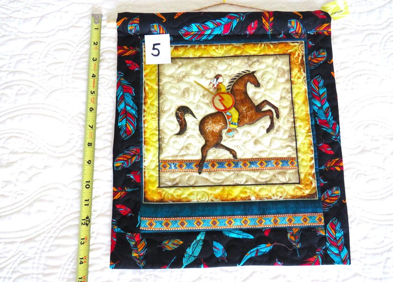 May include: Quilted wall hanging featuring a Native American on horseback. The central image is framed with a yellow border and a dark blue and turquoise border. The outer border is black with colorful feather designs. The wall hanging measures approximately 14 inches in length.