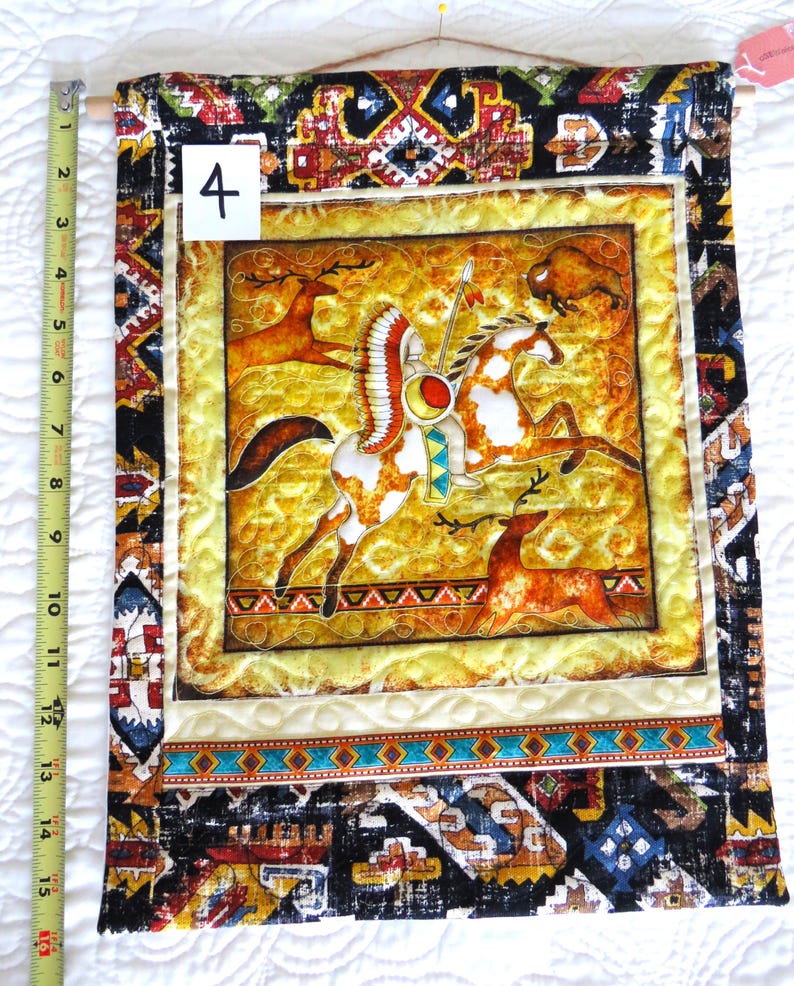 May include: A rectangular wall hanging with a Native American theme. The central image depicts a Native American on horseback with a buffalo and deer. The border features geometric patterns in black, brown, and turquoise. The wall hanging is approximately 16 inches long.
