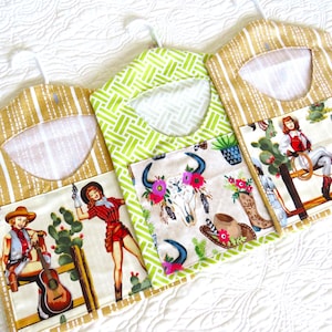 May include: Three fabric hanger covers with a Western theme. The covers feature a variety of patterns, including a tan and white stripe, a green geometric design, and a cream-colored fabric with cowgirl illustrations. Each cover has a pocket with a different cowgirl scene.