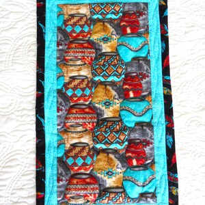 May include: A rectangular wall hanging featuring a repeating pattern of Southwestern pottery in shades of turquoise, red, brown, and gray. The central panel is framed by a turquoise border and a black border with a feather design. A wooden dowel is at the top.