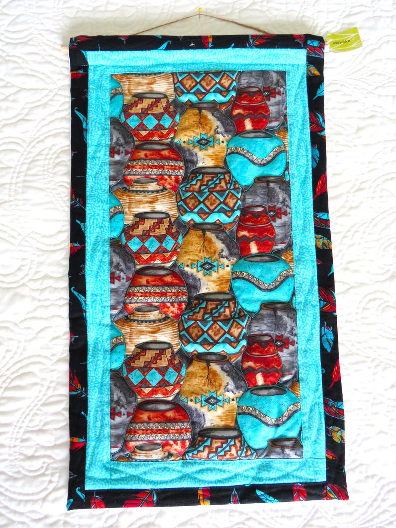 May include: A rectangular wall hanging featuring a repeating pattern of Southwestern pottery in shades of turquoise, red, brown, and gray. The central panel is framed by a turquoise border and a black border with a feather design. A wooden dowel is at the top.