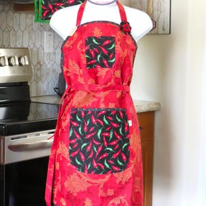 May include: A red apron with a gold paisley pattern, featuring a black pocket and a black hem, both decorated with a red and green chili pepper print. The apron has a black square chest pocket with the same chili print and a small red flower accent.