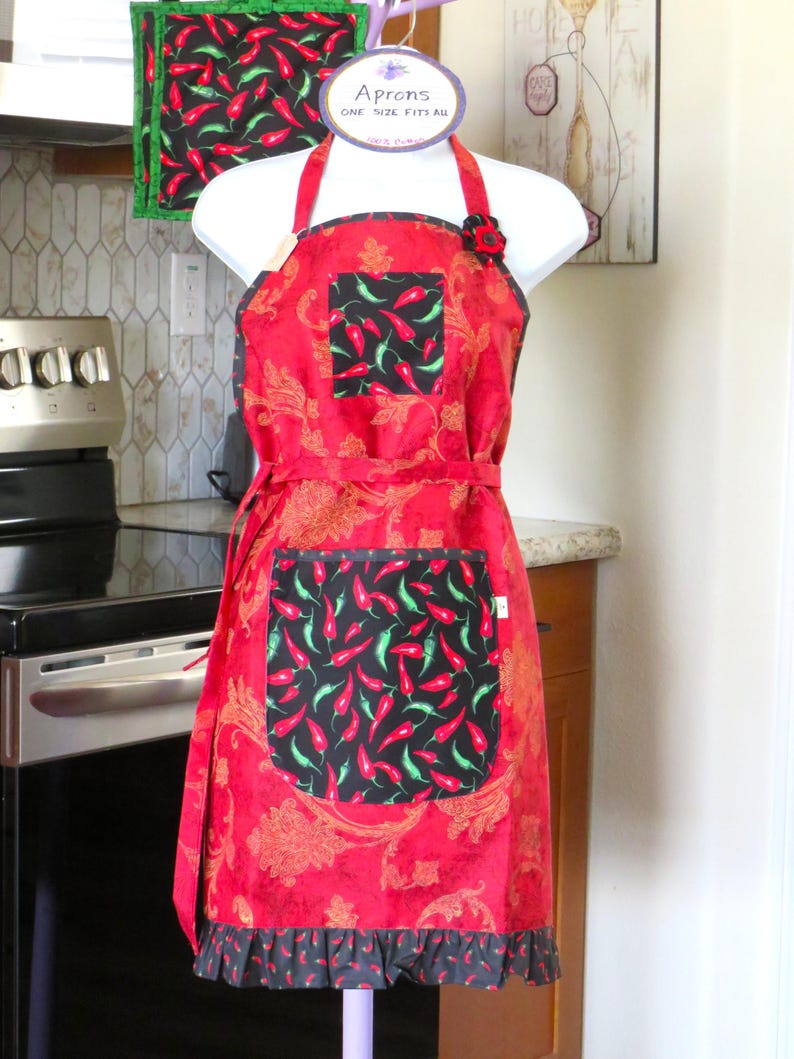 May include: A red apron with a gold paisley pattern, featuring a black pocket and a black hem, both decorated with a red and green chili pepper print. The apron has a black square chest pocket with the same chili print and a small red flower accent.