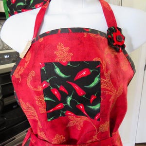 May include: A red apron featuring a gold paisley pattern and a central black square pocket decorated with a print of red and green chili peppers. The apron includes a fabric flower accent on the left strap with a dark brown button center.