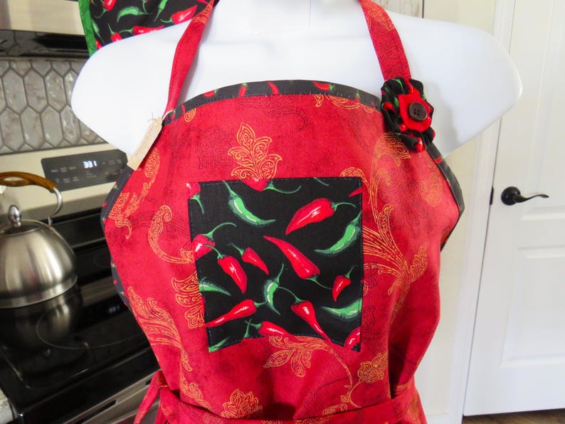 May include: A red apron featuring a gold paisley pattern and a central black square pocket decorated with a print of red and green chili peppers. The apron includes a fabric flower accent on the left strap with a dark brown button center.
