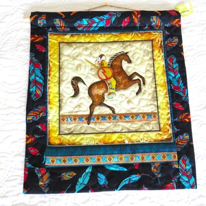 May include: Quilted wall hanging featuring a Native American rider on a brown horse. The central image is framed with a yellow border and surrounded by a black fabric adorned with colorful feather patterns. The artwork is a square shape.