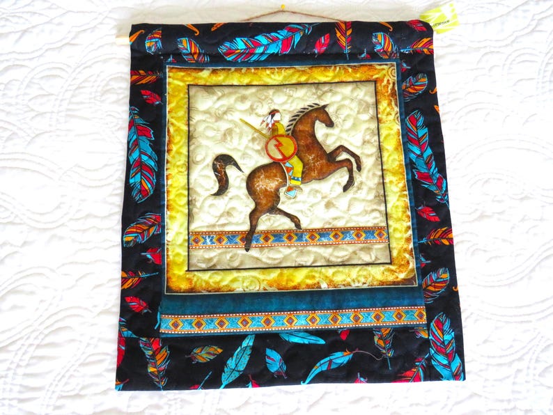 May include: Quilted wall hanging featuring a Native American rider on a brown horse. The central image is framed with a yellow border and surrounded by a black fabric adorned with colorful feather patterns. The artwork is a square shape.