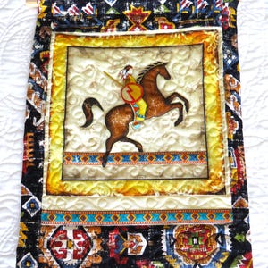 May include: A quilted wall hanging featuring a Native American on horseback. The central image is framed with a yellow border and surrounded by patterned fabric in black, red, and blue. A wooden dowel and pin are at the top.