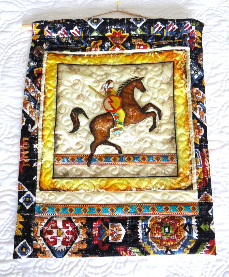 May include: A quilted wall hanging featuring a Native American on horseback. The central image is framed with a yellow border and surrounded by patterned fabric in black, red, and blue. A wooden dowel and pin are at the top.