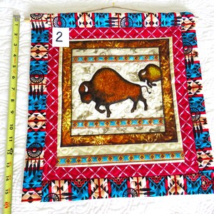 May include: A fabric wall hanging featuring a brown bison and calf design. The artwork is framed with a red and turquoise geometric pattern, and measures approximately 15 inches in length. The wall hanging is mounted on a wooden dowel.
