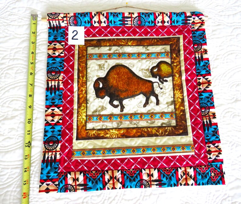 May include: A fabric wall hanging featuring a brown bison and calf design. The artwork is framed with a red and turquoise geometric pattern, and measures approximately 15 inches in length. The wall hanging is mounted on a wooden dowel.