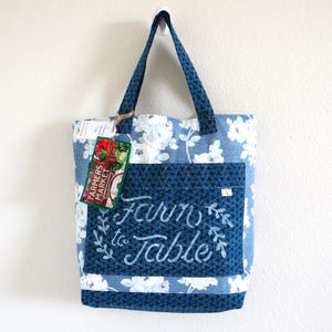 May include: A blue and white floral print tote bag with two handles. The bag features a dark blue front pocket with the embroidered white text Farm to Table and a decorative green tag labeled Farmers Market attached to the handle with twine.