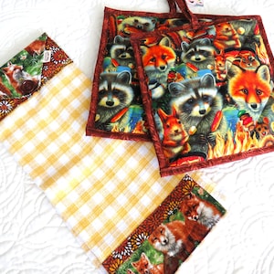 May include: Two red and brown quilted pot holders with a woodland animal print featuring raccoons and foxes. The pot holders are on a white and yellow checkered kitchen towel.