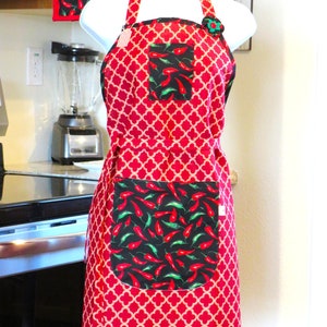 May include: A red and beige quatrefoil patterned apron featuring a black chest pocket and a large black lower pocket, both decorated with a vibrant print of red and green chili peppers. The apron includes a decorative fabric flower at the neckline.