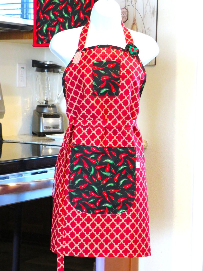 May include: A red and beige quatrefoil patterned apron featuring a black chest pocket and a large black lower pocket, both decorated with a vibrant print of red and green chili peppers. The apron includes a decorative fabric flower at the neckline.