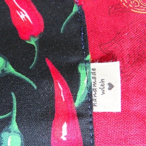 May include: A fabric item featuring a black section with a pattern of red and green chili peppers and a solid red section. A small white rectangular tag is sewn into the seam, displaying the text "handmade with" followed by a heart symbol.