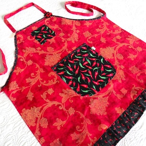 May include: A red apron featuring a gold floral damask pattern, accented with black pockets and a bottom ruffle decorated with a red and green chili pepper print. The apron includes matching red fabric neck and waist ties for securing the garment.