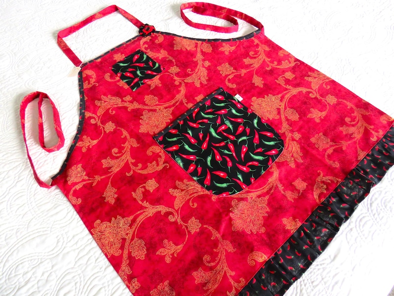 May include: A red apron featuring a gold floral damask pattern, accented with black pockets and a bottom ruffle decorated with a red and green chili pepper print. The apron includes matching red fabric neck and waist ties for securing the garment.