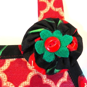 May include: A fabric apron featuring a red and beige geometric lattice pattern on the lower section and a black fabric with a red cherry print on the upper strap. A gathered black cherry-print fabric rosette with a green felt flower and a red button.