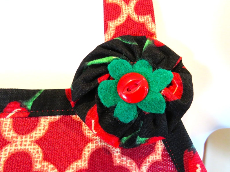 May include: A fabric apron featuring a red and beige geometric lattice pattern on the lower section and a black fabric with a red cherry print on the upper strap. A gathered black cherry-print fabric rosette with a green felt flower and a red button.
