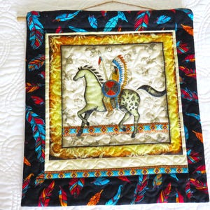 May include: Quilted wall hanging featuring a Native American on horseback. The central image is framed with a gold border and surrounded by a black fabric with colorful feathers. The artwork is mounted on a wooden dowel.