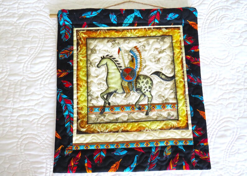 May include: Quilted wall hanging featuring a Native American on horseback. The central image is framed with a gold border and surrounded by a black fabric with colorful feathers. The artwork is mounted on a wooden dowel.