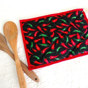 May include: Red and green chili pepper patterned hot pad with a red border. The hot pad is on a white surface with two wooden spoons.