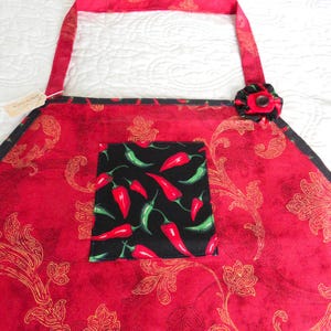 May include: A red apron featuring a gold paisley pattern, a black square pocket with a red and green chili pepper print, and a black fabric flower accent with a central button attached to the upper right corner of the apron's main body.