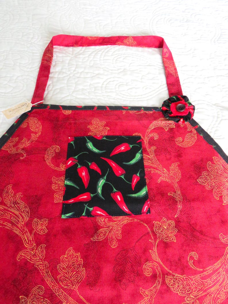May include: A red apron featuring a gold paisley pattern, a black square pocket with a red and green chili pepper print, and a black fabric flower accent with a central button attached to the upper right corner of the apron's main body.