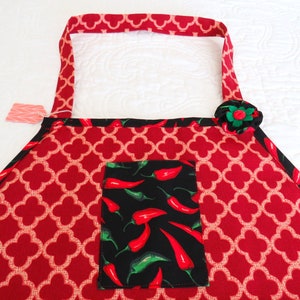 May include: A red apron featuring a white quatrefoil pattern, black trim, and a central black pocket decorated with a print of red and green chili peppers. A fabric flower in black, red, and green is attached to the upper right corner of the apron.