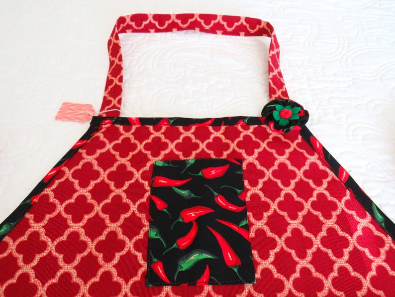May include: A red apron featuring a white quatrefoil pattern, black trim, and a central black pocket decorated with a print of red and green chili peppers. A fabric flower in black, red, and green is attached to the upper right corner of the apron.