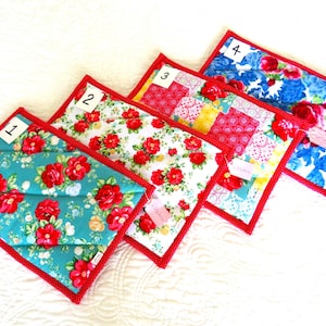 May include: Four quilted placemats with floral patterns and red trim. The placemats feature different floral designs, including red roses on turquoise, white, and blue backgrounds. Each placemat has a red border and is numbered.