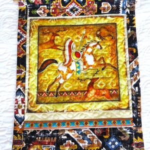 May include: A decorative wall hanging featuring a Native American warrior on horseback, with deer and a bison. The central image is framed by a yellow border and a patterned fabric border with geometric designs in black, brown, and blue. The hanging is attached to a wooden dowel.