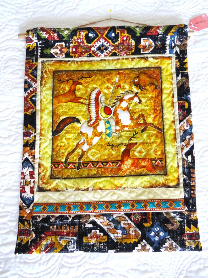 May include: A decorative wall hanging featuring a Native American warrior on horseback, with deer and a bison. The central image is framed by a yellow border and a patterned fabric border with geometric designs in black, brown, and blue. The hanging is attached to a wooden dowel.