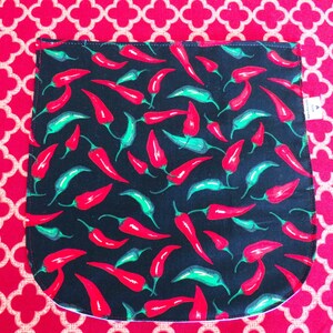 May include: A black fabric pouch featuring a repeating pattern of red and green chili peppers. The pouch has a rounded bottom edge and a small white tag on the right side. The fabric is displayed against a bright red background with a white quatrefoil pattern.
