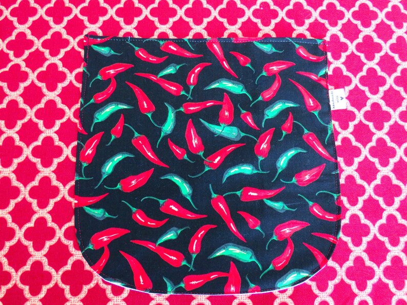 May include: A black fabric pouch featuring a repeating pattern of red and green chili peppers. The pouch has a rounded bottom edge and a small white tag on the right side. The fabric is displayed against a bright red background with a white quatrefoil pattern.