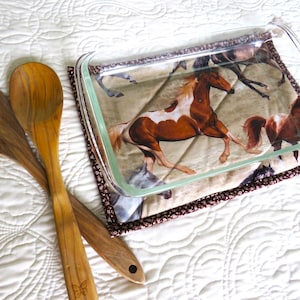 May include: A glass baking dish with a horse print design on a brown quilted hot pad. Two wooden spoons are on a white quilted surface.