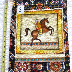 May include: A quilted wall hanging featuring a Native American on horseback. The central image is framed with a yellow border and surrounded by patterned fabric. The hanging is approximately 15 inches long, with the number "3" visible.