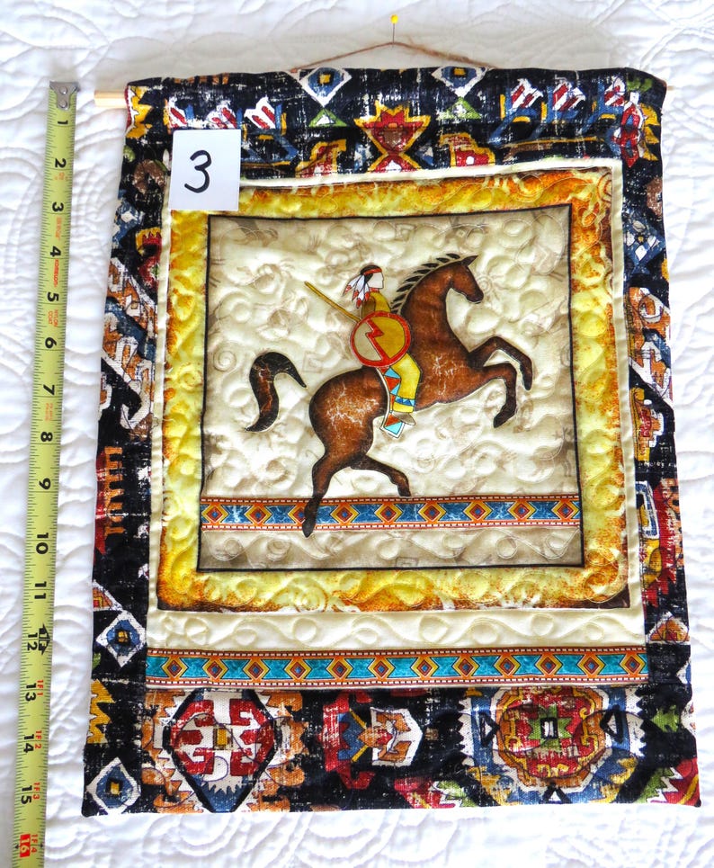 May include: A quilted wall hanging featuring a Native American on horseback. The central image is framed with a yellow border and surrounded by patterned fabric. The hanging is approximately 15 inches long, with the number "3" visible.