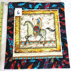 May include: Quilted wall hanging featuring an Native American on horseback. The central image is framed with a gold border and a black border with colorful feathers. The number "6" is visible in the upper left corner. The hanging is approximately 14 inches long.