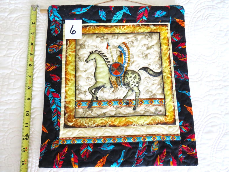 May include: Quilted wall hanging featuring an Native American on horseback. The central image is framed with a gold border and a black border with colorful feathers. The number "6" is visible in the upper left corner. The hanging is approximately 14 inches long.