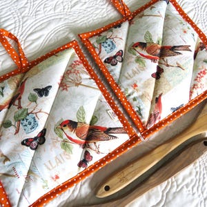 May include: Two quilted pot holders with a bird and butterfly print, trimmed with orange polka dot fabric. The pot holders have orange hanging loops. Wooden spoons are next to the pot holders.