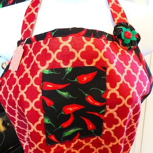 May include: A red apron featuring a repeating beige quatrefoil pattern, accented with a black pocket displaying a vibrant red and green chili pepper print. The apron includes a decorative fabric flower on the strap and a small paper tag.