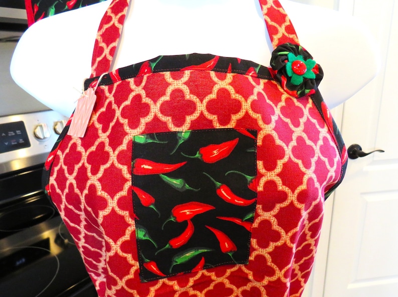 May include: A red apron featuring a repeating beige quatrefoil pattern, accented with a black pocket displaying a vibrant red and green chili pepper print. The apron includes a decorative fabric flower on the strap and a small paper tag.