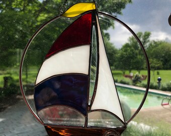 May include: A stained glass sailboat suncatcher with a red, white and blue sail, a yellow sail at the top, and a brown hull. The suncatcher is hanging from a window with a chain and a suction cup.