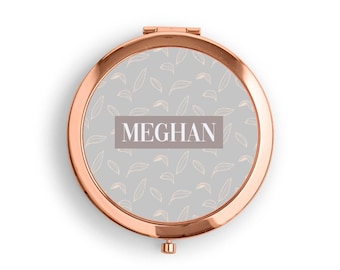 May include: A rose gold compact mirror with a gray background and a white leaf pattern. The name "MEGHAN" is printed in white on the mirror.