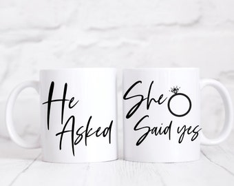 May include: Two white ceramic mugs with black text. The mug on the left says "He Asked" and the mug on the right says "She Said Yes" with a simple ring and diamond graphic.
