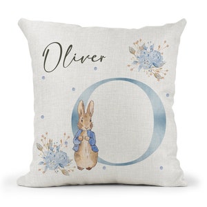 May include: A white pillow with a blue and brown illustration of Peter Rabbit, the letter O, and the name Oliver. The pillow has a light blue and brown floral design.